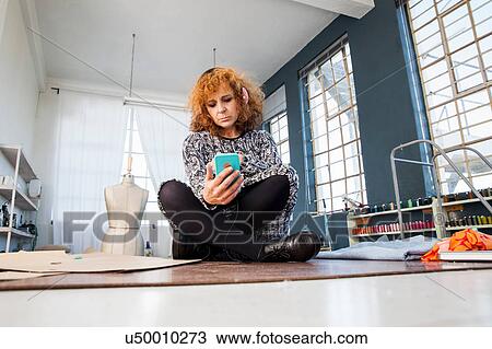 Stock Image - Mature female fashion designer cross-legged on workshop table looking at smartphone. Fotosearch