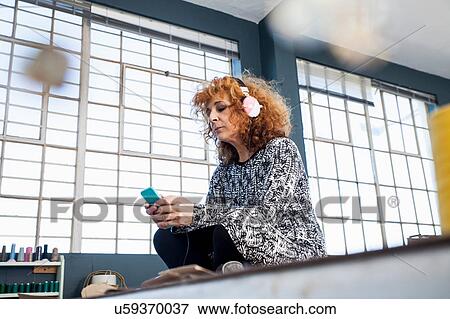 Stock Photo - Mature female fashion designer listening to smartphone music in workshop. Fotosearch
