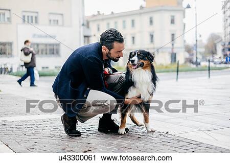 Stock Image - Mid adult man crouching to pet dog in cobbled city square. Fotosearch