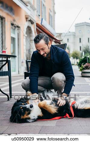 Stock Photo - Mid adult man crouching to pet dog in city square. Fotosearch