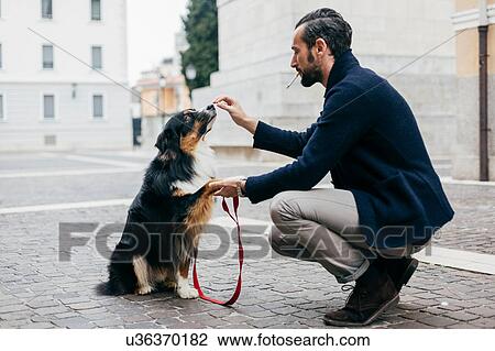 Stock Image - Mid adult man crouching with his dog in cobbled city square. Fotosearch