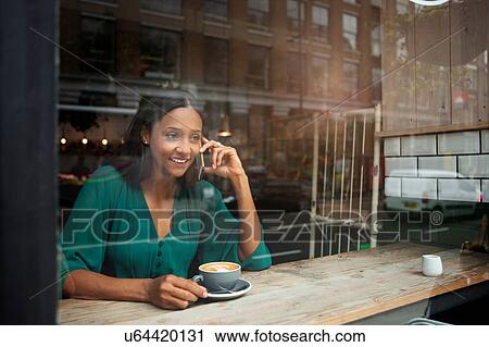 Stock Image - Mid adult woman chatting on smartphone in cafe window seat. Fotosearch