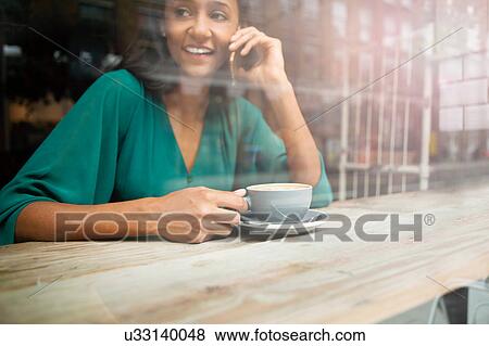 Stock Photo - Mid adult woman chatting on smartphone in cafe window seat. Fotosearch