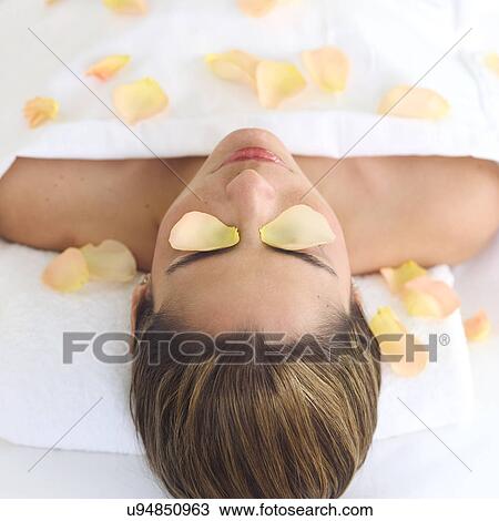 Stock Image - Mid adult woman lying on back under sheet covered with rose petals. Fotosearch