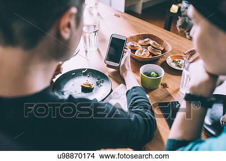 Picture - Over shoulder view of two young female friends reading smartphone texts at kitchen table. Fotosearch