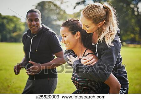 Personal trainer encouraging two women piggy backing in park View Large Photo Image Stock Photo - Personal trainer encouraging two women piggy backing in park. Fotosearch