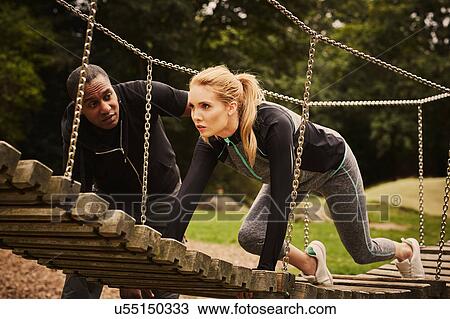 Stock Image - Personal trainer instructing young woman crawling on playground equipment in park. Fotosearch