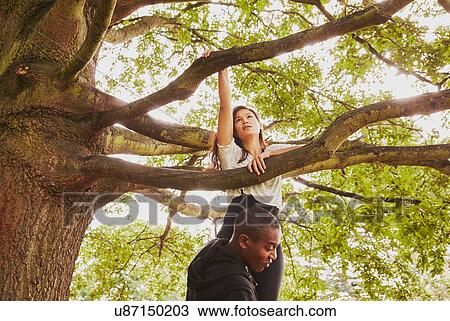Stock Image - Personal trainer lifting woman to climb park tree. Fotosearch