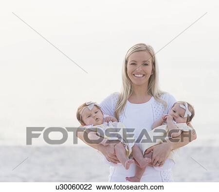 Stock Image - Portrait of happy mid adult mother carrying two baby girls (2-5 months). Fotosearch
