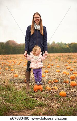 Stock Image - Portrait of mid adult woman and toddler daughter standing on top of pumpkin in field. Fotosearch