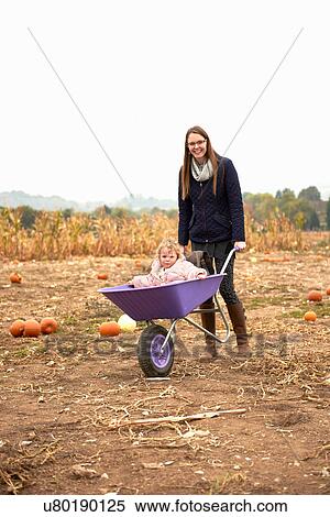 Stock Photography - Portrait of mid adult woman pushing toddler daughter in wheelbarrow in pumpkin field. Fotosearch