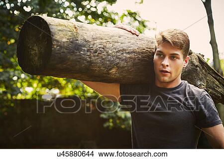 Picture - Portrait of young man training, lifting tree trunk on shoulder in park. Fotosearch