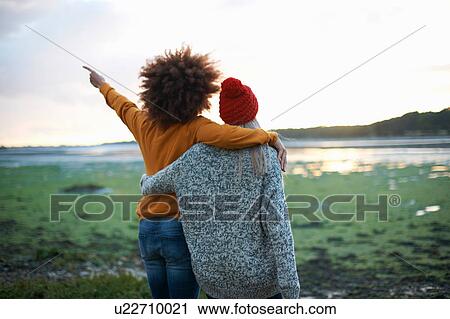 Stock Image - Rear view of two young women by sea pointing at sunset. Fotosearch