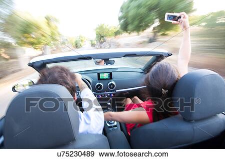 Rear view of young couple taking selfie whilst driving on rural road in convertible, Majorca, Spain View Large Photo Image Stock Photo - Rear view of young couple taking selfie whilst driving on rural road in convertible, Majorca, Spain. Fotosearch
