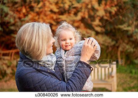 Stock Photograph - Senior woman carrying toddler granddaughter in autumn park. Fotosearch