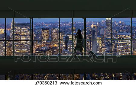 Stock Image - Silhouetted woman crossing city footbridge over New York cityscape at night, USA. Fotosearch
