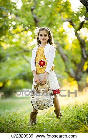 Stock Photo - Smiling girl standing in orchard, holding basket with teddy bear. Fotosearch