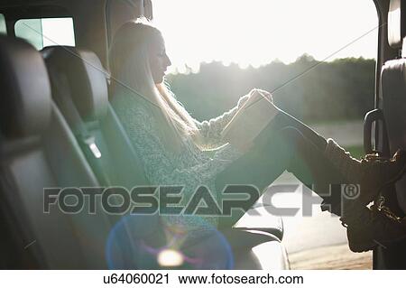 Stock Image - Sunlit portrait of young woman reading book at back seat of car. Fotosearch