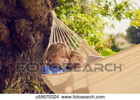 Picture - Teenage boy and brother reclining in garden hammock browsing digital tablet. Fotosearch