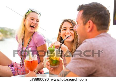 Stock Image - Three adult friends laughing over cocktails at waterfront restaurant, Majorca, Spain. Fotosearch