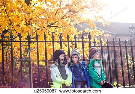 Stock Photo - Three boys, outdoors, leaning against fence, in autumn. Fotosearch