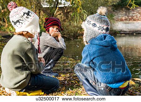 Stock Photo - Three young boys, outdoors, crouching beside lake. Fotosearch