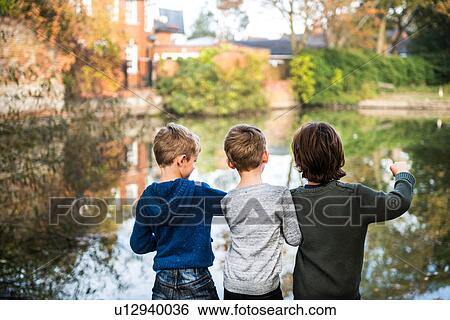 Stock Photograph - Three young boys, standing beside lake, rear view. Fotosearch