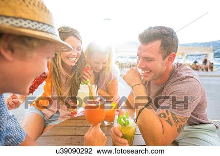 Stock Image - Two adult couples laughing over cocktails at waterfront restaurant, Majorca, Spain. Fotosearch