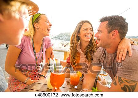 Stock Image - Two adult couples laughing over cocktails at waterfront restaurant, Majorca, Spain. Fotosearch