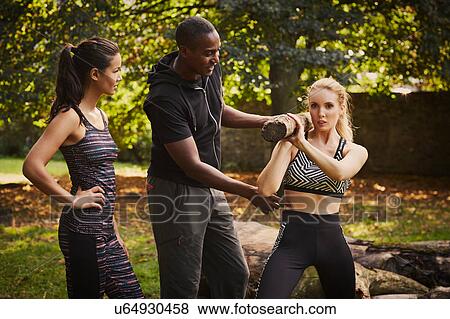 Stock Photo - Two women with personal trainer explaining tree trunk shoulder lift in park. Fotosearch