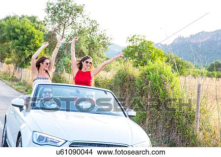 Two young female friends standing waving whilst driving on rural road in convertible, Majorca, Spain View Large Photo Image Picture - Two young female friends standing waving whilst driving on rural road in convertible, Majorca, Spain. Fotosearch