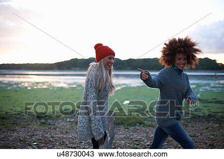 Stock Image - Two young women laughing by sea at sunset. Fotosearch