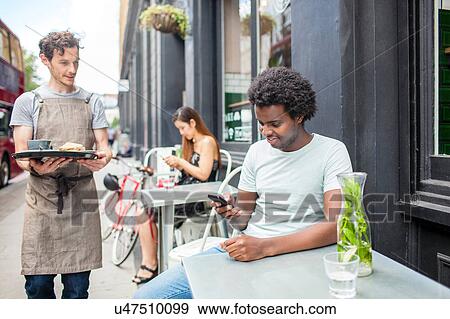 Waiter serving young man looking at smartphone at city sidewalk cafe View Large Photo Image Stock Photo - Waiter serving young man looking at smartphone at city sidewalk cafe. Fotosearch