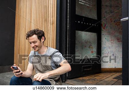 Stock Photo - Waiter taking a break on cafe step looking at smartphone. Fotosearch