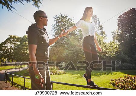 Stock Photo - Woman holding hands with personal trainer walking on park fence. Fotosearch