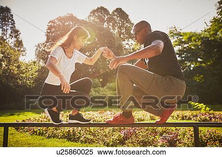 Stock Photography - Woman holding hands with personal trainer crouching and balancing on park fence. Fotosearch