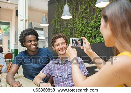 Stock Photo - Woman photographing male friends on smartphone at cafe table. Fotosearch