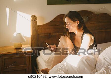 Stock Image - Woman using mobile phone in bed. Fotosearch