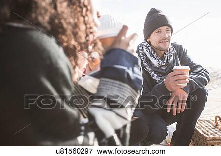 Stock Photo - Young adult friends crouching on beach with takeaway coffee, Western Cape, South Africa. Fotosearch