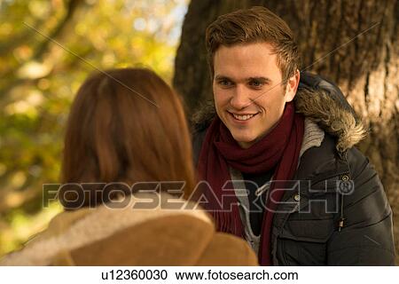 Stock Image - Young couple in forest, sitting face to face, smiling. Fotosearch