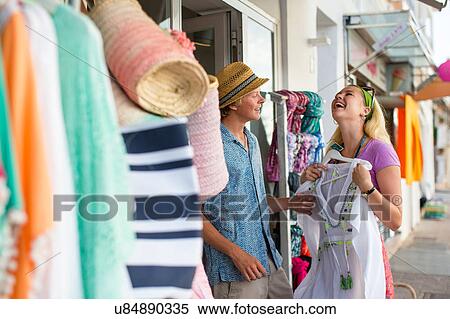 Stock Photography - Young couple laughing trying sundress at shop front, Majorca, Spain. Fotosearch