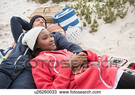 Stock Image - Young couple reclining on picnic blanket at beach, Western Cape, South Africa. Fotosearch