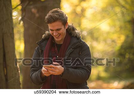 Picture - Young man in forest, looking at smartphone, smiling. Fotosearch