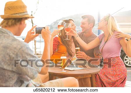 Stock Image - Young man photographing adult friends at waterfront restaurant, Majorca, Spain. Fotosearch