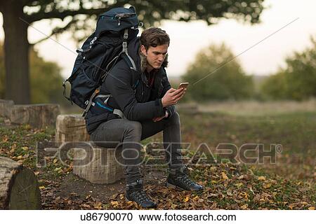 Stock Image - Young man sitting on tree stump, using smartphone. Fotosearch