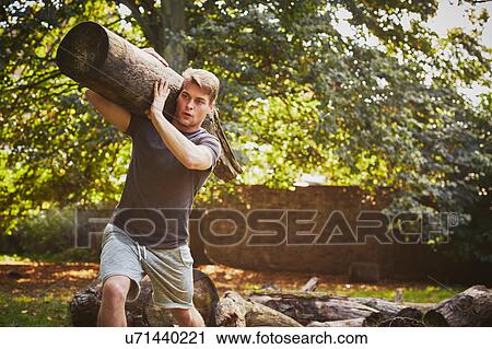 Stock Image - Young man training, lifting tree trunk on shoulder in park. Fotosearch