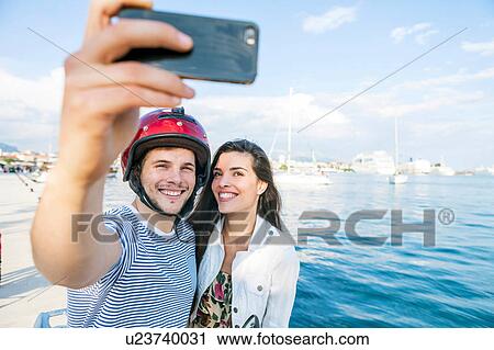 Stock Image - Young moped couple taking selfie on harbour, Split, Dalmatia, Croatia. Fotosearch