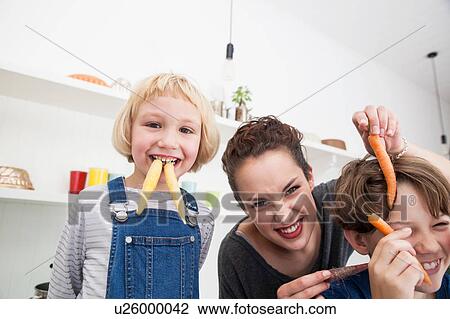 Stock Image - Young woman, boy and girl in kitchen, fooling around, using carrots as false teeth. Fotosearch