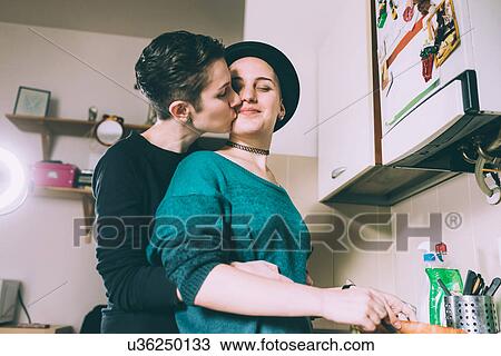 Stock Image - Young woman kissing girlfriend in kitchen. Fotosearch
