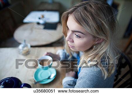 Stock Image - Young woman sitting in cafe, holding tea cup, thoughtful expression on face. Fotosearch